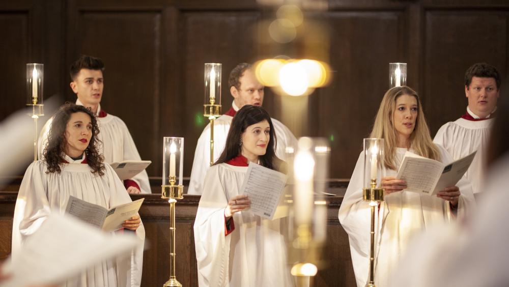 A group of choir singers in white robes stand in wooden choir stalls holding sheet music. Tall candle holders with lit candles are positioned in front of them. The singers appear focused and engaged in performance within a traditional chapel setting.