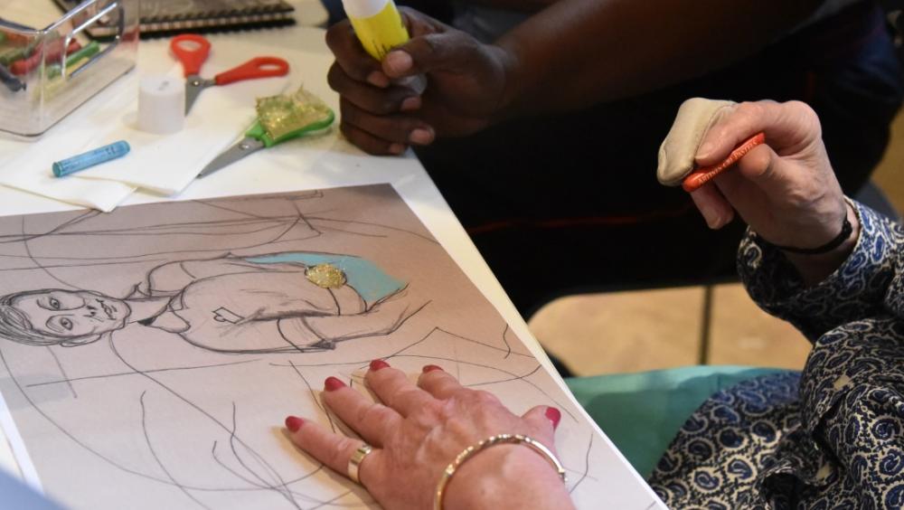 Hands of several people are shown holding various art implements, including crayons & glue. On a table are craft supplies and a sheet of paper depicting a hand-drawn sketch of a nurse.