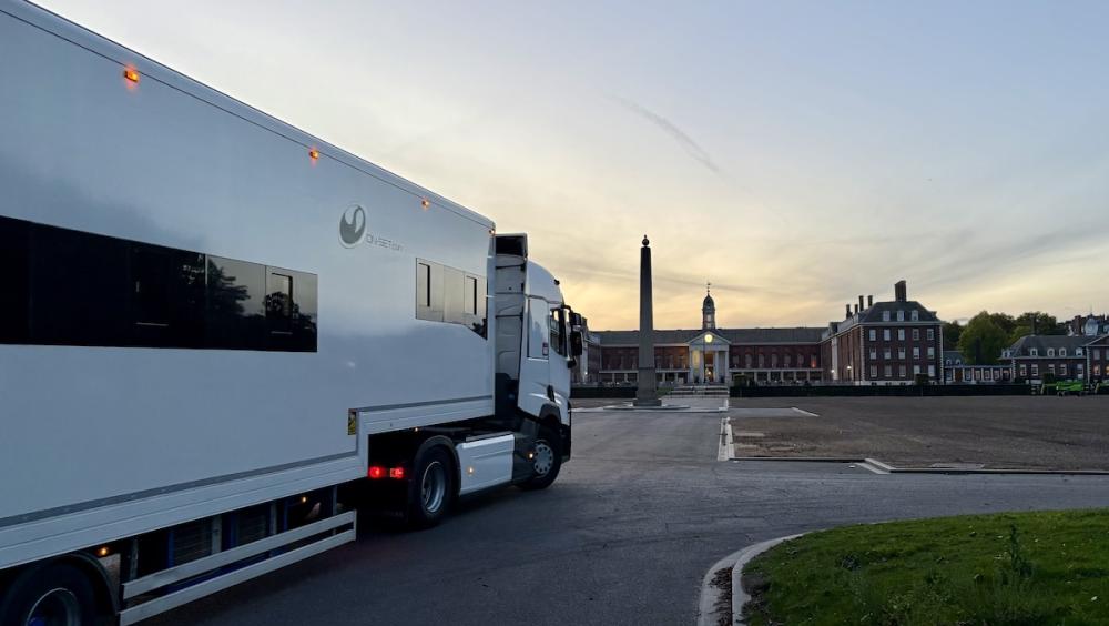 A large white production truck is parked at dawn near the grounds of the Royal Hospital Chelsea. The sky is pale with early morning light. In the distance, the Main Building and obelisk monument are visible. The scene is quiet, with empty paved areas and trimmed grass framing the approach to the historic buildings.