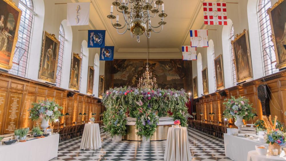 A grand historic hall with black and white chequered flooring, tall arched windows, and wood panelled walls engraved with names. Large portraits in gilded frames line both sides of the room. Colourful heraldic banners hang from the ceiling. In the centre stands an elaborate floral installation with cascading greenery and vibrant flowers surrounding a candelabra. Several round cocktail tables draped in white cloth are arranged around the space, along with buffet tables decorated with floral arrangements. A l