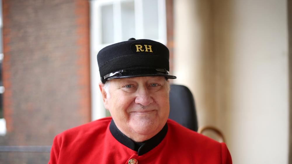 A Chelsea Pensioner wearing the traditional scarlet uniform and black cap smiles at the camera. He has several service medals displayed on his chest. The background shows the exterior of the Royal Hospital Chelsea with brickwork and white pillars softly out of focus.