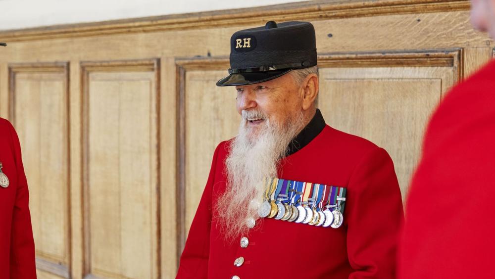 A group of Chelsea Pensioners stand in front of a wooden panelled wall, wearing their iconic scarlet uniforms with black Shako Hat emblazoned with gold "RH" lettering. The Pensioner in the centre has a long white beard and is highly decorated with almost a dozen medals.