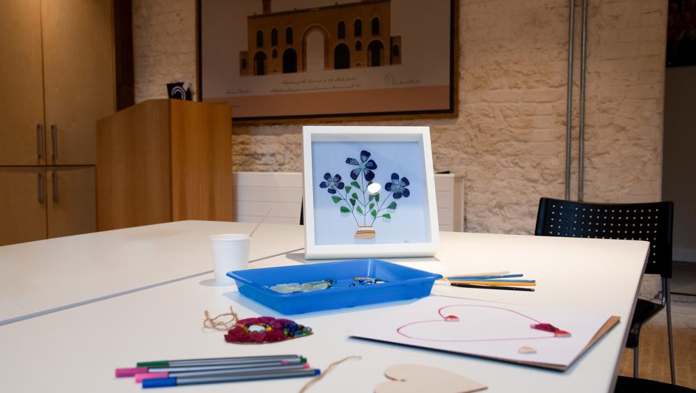 A well-lit craft room with a white table laid out for an arts activity. In the centre is a framed artwork of a flower bouquet made from paper cut-outs. Surrounding it are coloured pens, string, decorative gems, wooden shapes, and a drawing of a heart in progress. A textured brick wall and architectural print provide a warm backdrop.