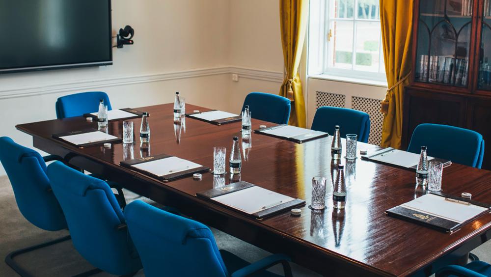 A polished wooden boardroom table surrounded by modern blue chairs, each place set with a notepad, pen, water bottle, and glass. A large flat-screen monitor is mounted on the wall, and sunlight streams through a window with gold curtains. A traditional wooden bookcase filled with books sits in the corner, blending modern meeting facilities with classic elegance.