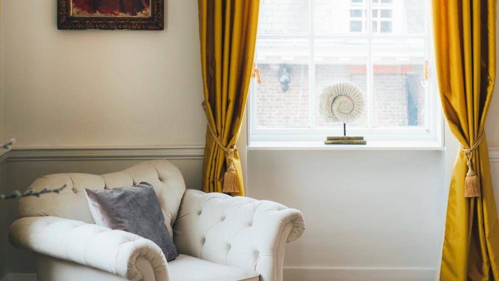 A cosy corner of a refined room featuring a tufted cream armchair with a grey cushion, placed beside a tall window framed by mustard yellow curtains. On the wall hangs a portrait of a Chelsea Pensioner in traditional uniform. A decorative ammonite fossil is displayed on the windowsill, with soft natural light illuminating the space.
