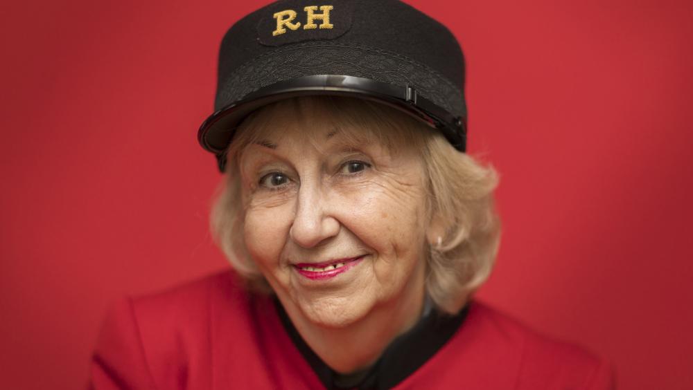 Portrait of a smiling Chelsea Pensioner wearing a scarlet uniform and black RH cap against a red background.