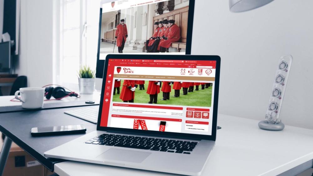 A laptop screen and desktop monitor are open and displaying various Chelsea Pensioner imagery, set upon a grey clean minimalist desk