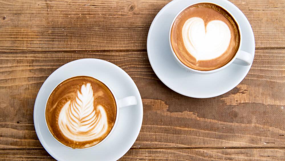 Two cups of coffee in white cups with saucers, upon a wooden table. Birds Eye view depicting the latte art of a heart and a tulip