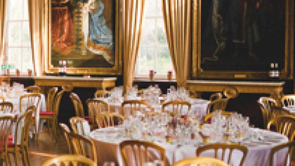 The State Apartments at The Royal Hospital Chelsea set out for a fine dining occasion with round tables and chairs, white table cloths and glassware arranged on each setting. Two large Van Dyke paintings hang on the walls in the background flanked by large portrait windows and opulent curtains