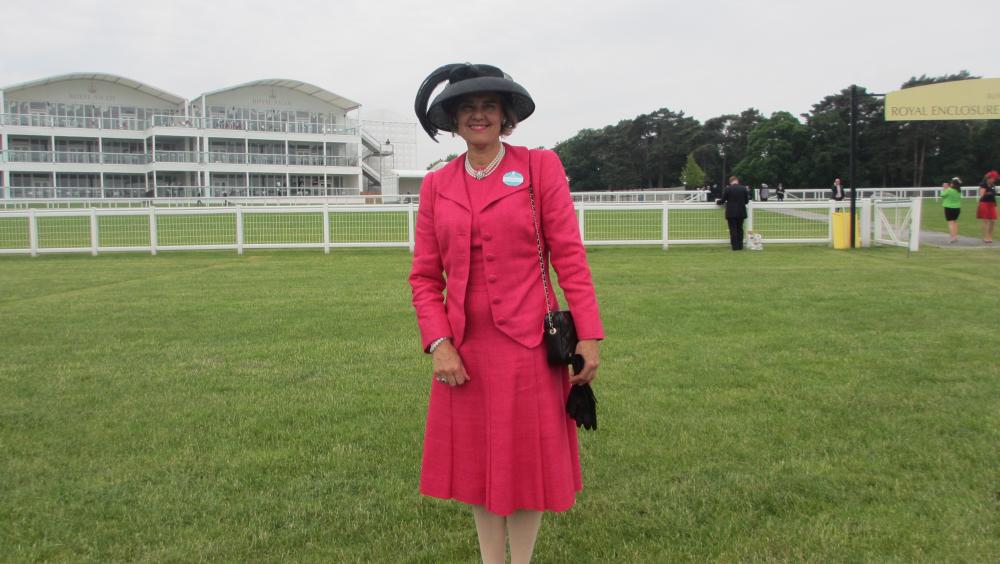A lady stands on the grounds of a horse racing track, dressed in a bright pink dress suit and black hat and holding a black pair of gloves and handbag.