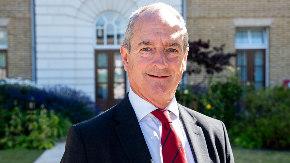 Portrait of The Royal Hospital Chelsea CEO, David Richmond, in a dark suit and red striped tie standing outdoors in sunlight, with a building and garden softly blurred behind him.