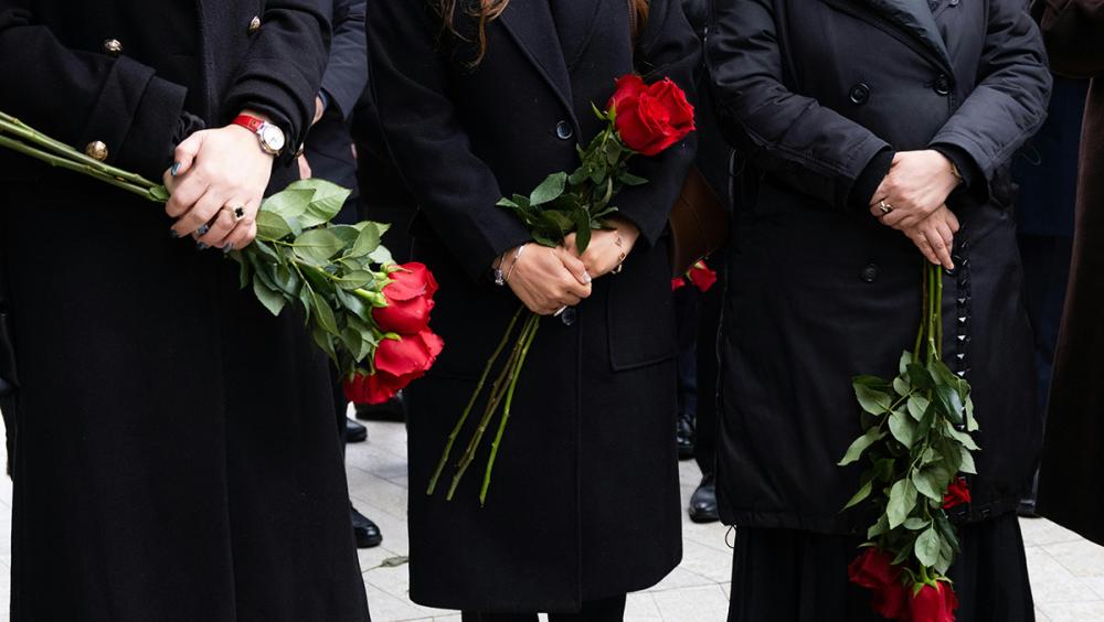 Close-up of two people in dark clothing holding red roses, suggesting a funeral or remembrance setting.