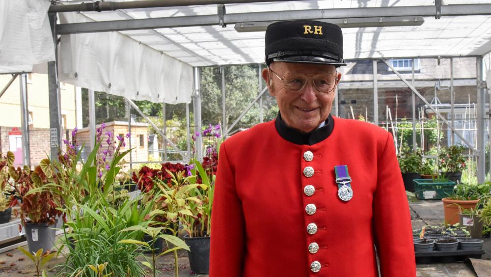 A Chelsea Pensioner in scarlet uniform stands inside a greenhouse surrounded by potted plants at the Royal Hospital Chelsea.