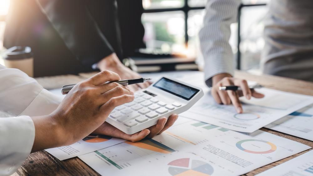 A group of finance staff sit around a table of reports and charts, while another uses a calculator