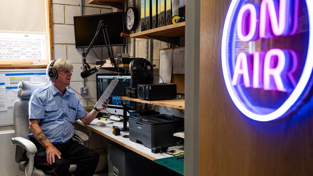 A Chelsea Pensioner in his blue shift, headphones on and script in hand, presenting from the on-air studio at the Royal Hospital Chelsea, with the illuminated "ON AIR" sign in the foreground.
