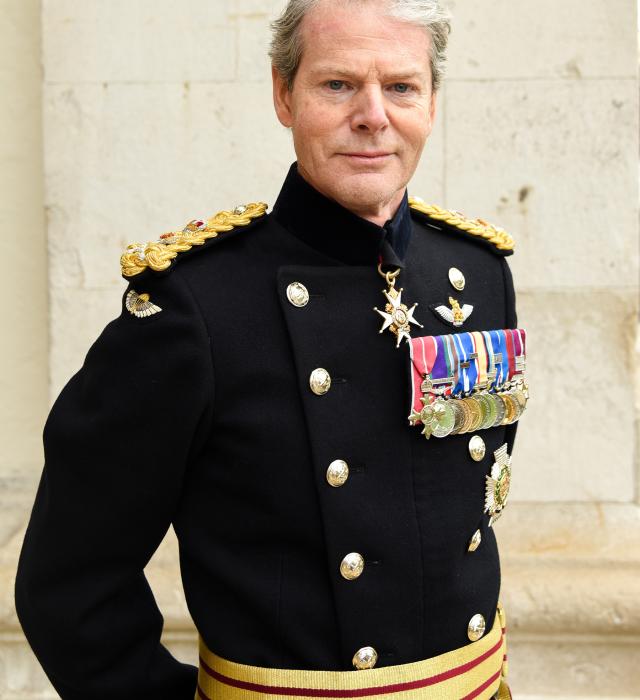 A highly decorated 4-star General of the British Army stands with hands behind his back against a white brick building