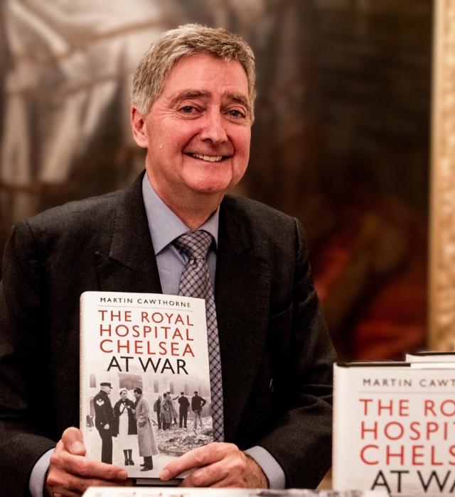 Author Martin Cawthorne is pictured wearing a suit and tie, holding a copy of his Royal Hospital Chelsea at War book, in front of a large portrait of Charles II within the State Apartments.