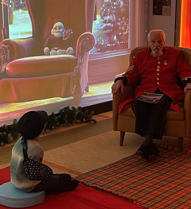 A Chelsea Pensioner sits in his iconic Scarlet uniform in a Christmas themed grotto, with a festive digital back drop. In front a young child with an arm in a sling listens carefully to a story
