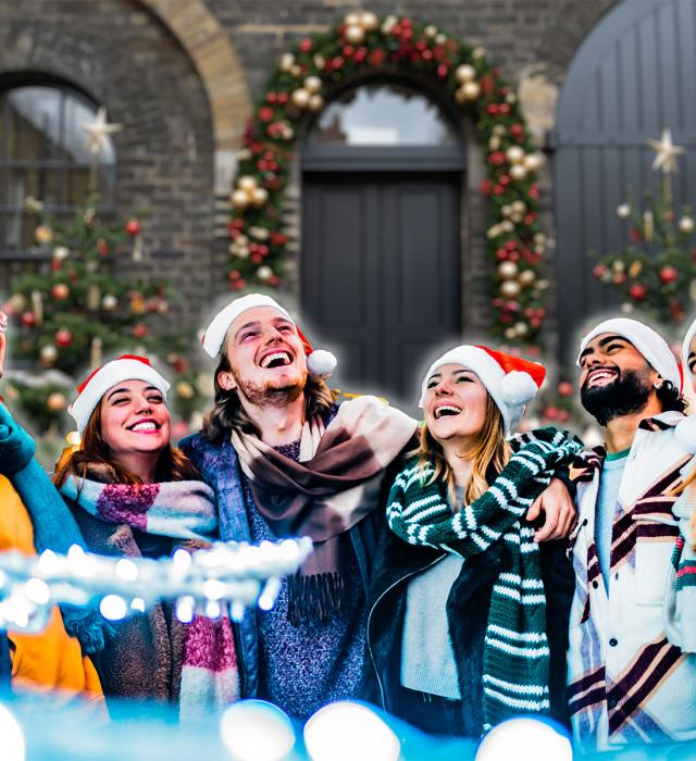 a group of friends dressed in Christmas hats and winter coats are laughing together in front of a festive decorated Soane Stable Courtyard