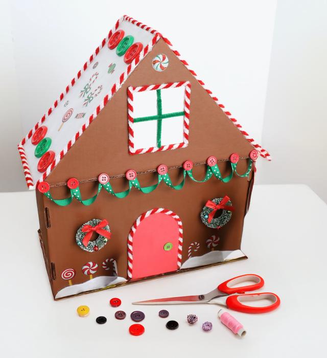 A cardboard gingerbread house sits atop a white table with colourful Christmas buttons and ribbons decorating the house. On the table are strewn buttons of various colours and a red pair of scissors