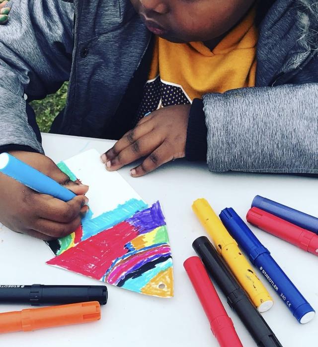 A child dressed in grey jacket and yellow t-shirt, holds a blue pen while colouring in a paper tree in multiple colours. Various coloured pens are strewn across the white desk.