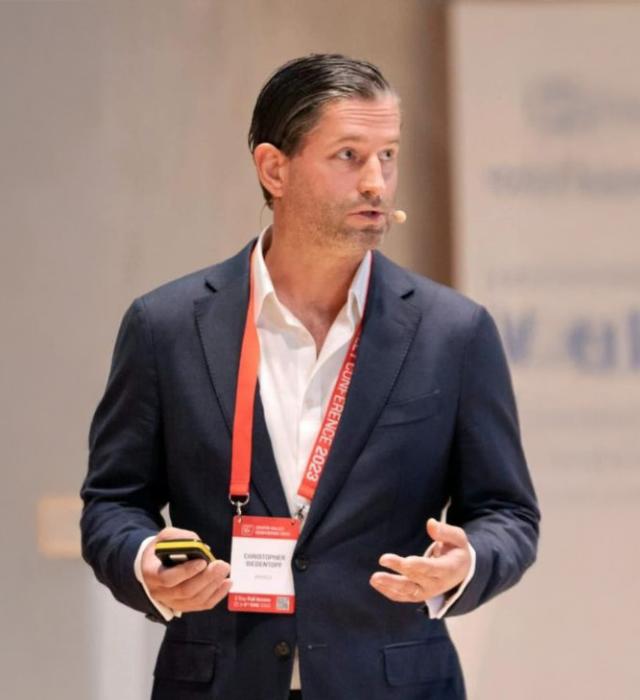 A suited guest speaker, Christopher Siedentopf, is shown talking, wearing a microphone and holding a remote control. A Red lanyard hangs around his neck.