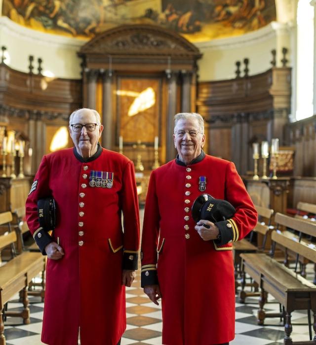 Two Chelsea Pensioners in traditional scarlet uniforms stand side by side inside the Wren Chapel at the Royal Hospital Chelsea. Wooden benches line the chapel, and the ornate altar and painted apse are visible behind them.