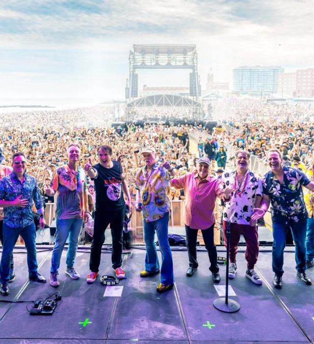 A large band stands across a festival stage facing a vast seaside crowd, smiling and gesturing towards the audience. Thousands of people fill the beach and promenade beyond, with a large outdoor stage structure and coastal buildings visible in the distance under a bright sky.