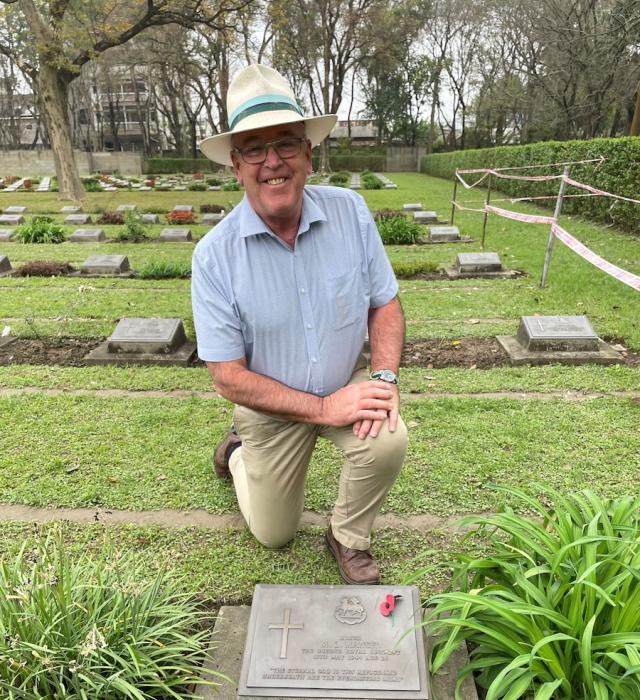 A man wearing a blue short sleeved shirt, cream trousers and a white Panama hat, kneels behind a war grave adorned with a single red poppy.