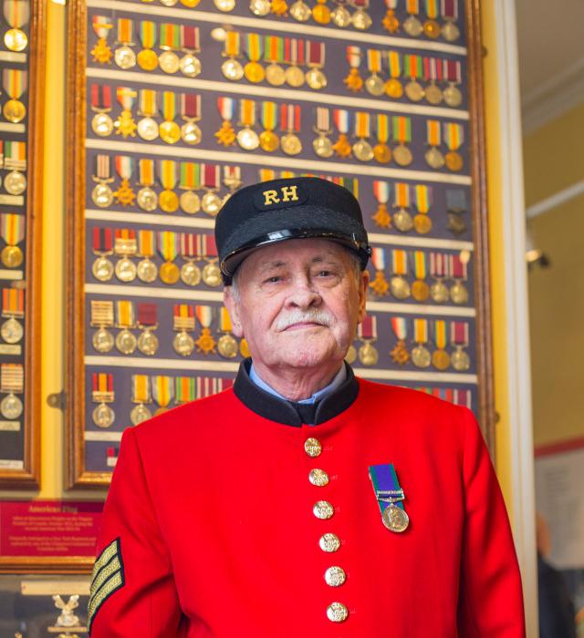 A Chelsea Pensioner stands in front of a wall of medals, wearing his iconic scarlet uniform adorned with a single medal on the lapel.