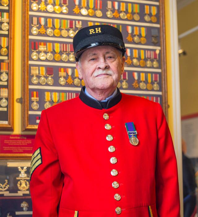 A Chelsea Pensioner stands in front of a wall of medals, wearing his iconic scarlet uniform adorned with a single medal on the lapel.