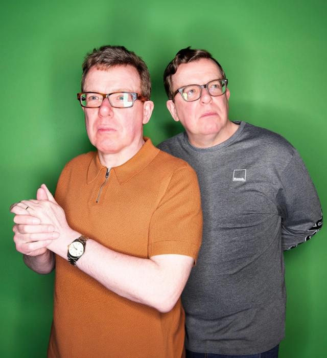 Two middle-aged men stand against a bright green background, both wearing glasses. One wears a short-sleeved rust-coloured polo shirt and clasps his hands, while the other stands slightly behind in a grey long-sleeved top. Both look off to the side with thoughtful, slightly serious expressions.