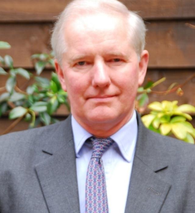 A man with grey hair stands in front of a wooden shed with yellow flowers and green foliage behind him. He wears a grey suit jacket, light shirt and purple patterned tie.