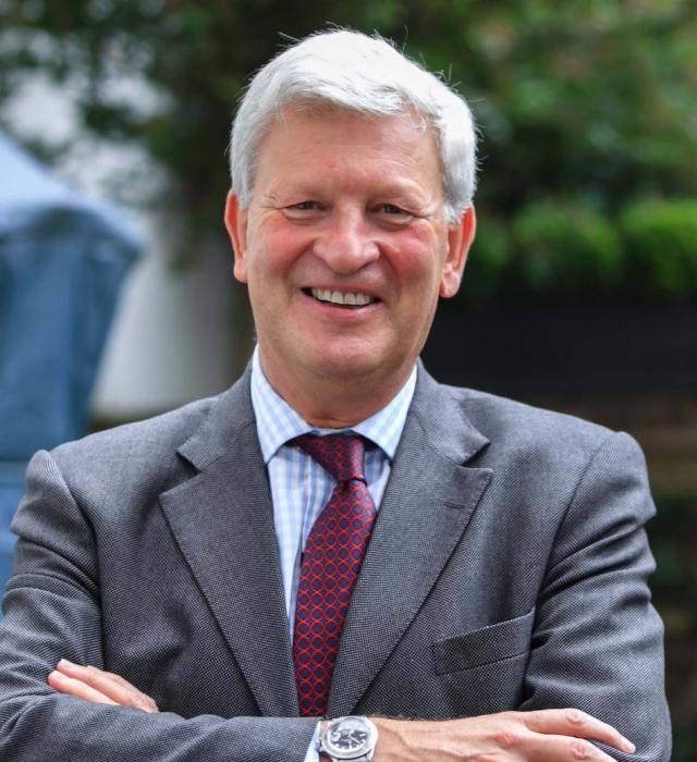 Sir Simon Mayall, a man stands with his arms crossed and a large welcoming smile on his face outdoors. His arms are folded in front of him and wears a grey suit with burgundy tie.