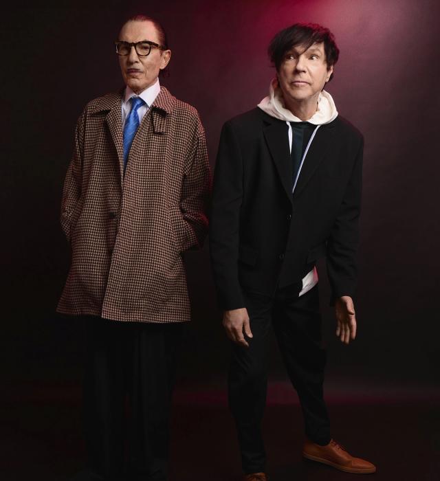 Two men stand side by side against a dark studio backdrop with subtle red lighting. One wears a patterned overcoat, glasses, and a tie, looking down thoughtfully, while the other wears a black suit layered over a light hooded top, looking off to the side. The image has a stylised, dramatic promotional feel.