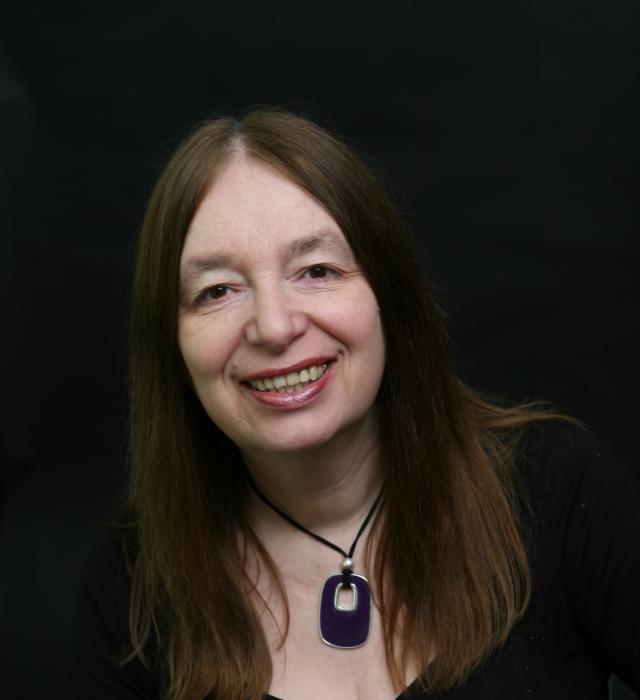 Portrait of a smiling woman with long brown hair wearing a black top and a purple pendant necklace, photographed against a dark background.