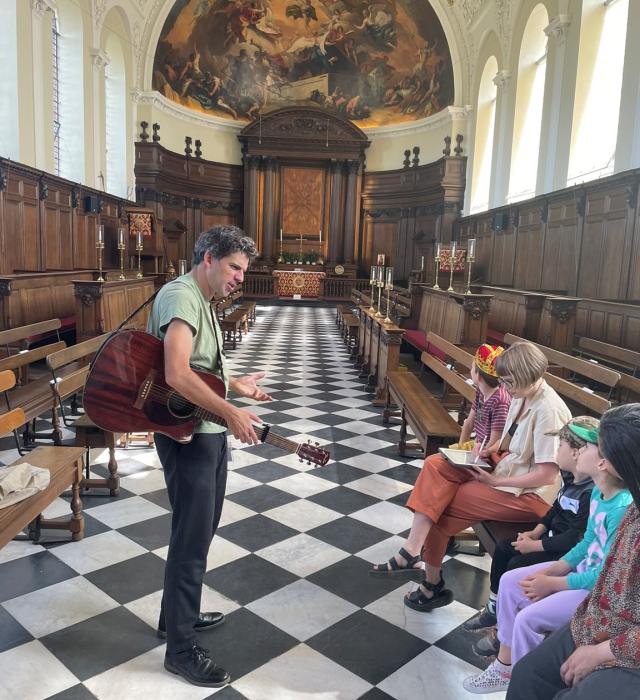 A man holding an acoustic guitar speaks to a small group of children seated on wooden pews inside an ornate chapel. The chapel features a black-and-white tiled floor, carved wooden panelling, and a large painted dome above the altar. The children listen attentively, suggesting an interactive family tour or musical storytelling session.