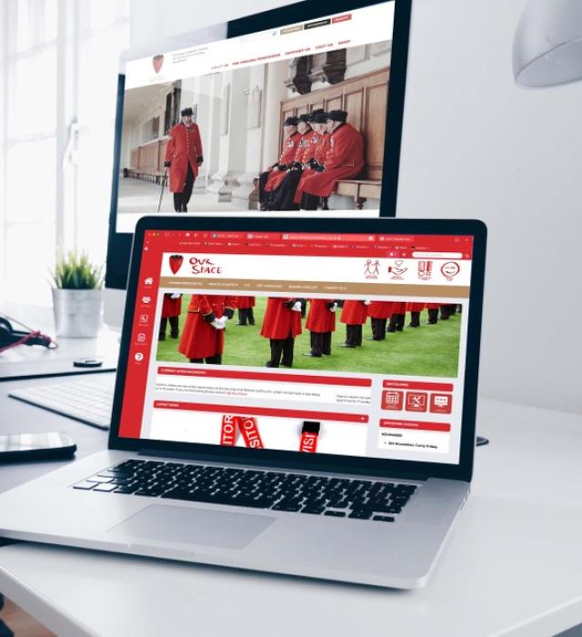 A laptop screen and desktop monitor are open and displaying various Chelsea Pensioner imagery, set upon a grey clean minimalist desk