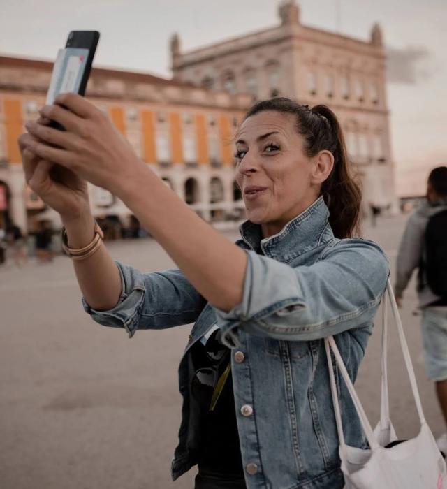 A woman wearing a denim jacket smiles while holding up a smartphone to take a photo or video in a large public square. Historic buildings with arches form the background, and other people are visible nearby, suggesting a lively city setting.