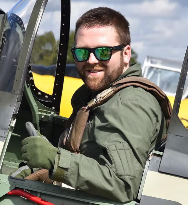 A man with short dark hair, sits within the cockpit of a WWII fighter jet, wearing Air Force flying wear and giving a thumbs up.