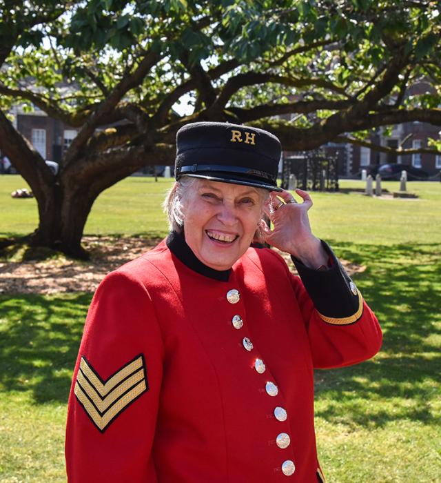 A smiling Chelsea Pensioner in scarlet uniform stands in the grounds of the Royal Hospital Chelsea beneath a tree.