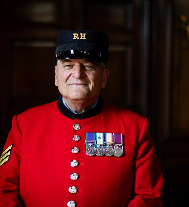 A Chelsea Pensioner stands in a wooden panelled room with his iconic scarlet uniform and a chest full of medals.