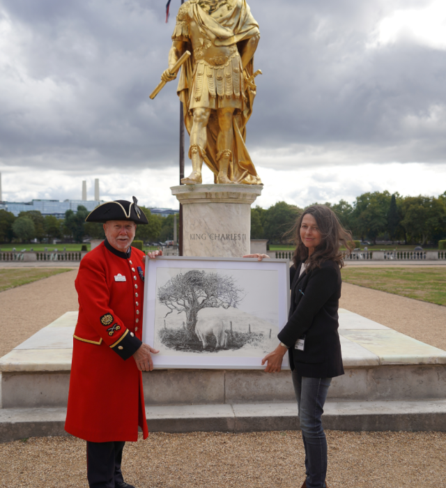 An image of Chelsea Pensioner Roy Palmer holding up a painting alongside a woman with a gold statue in the background at Royal Hospital Chelsea