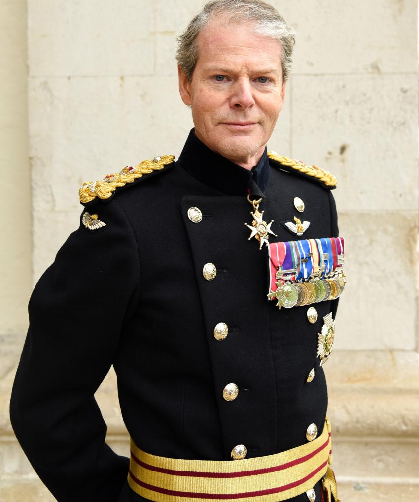 A highly decorated 4-star General of the British Army stands with hands behind his back against a white brick building