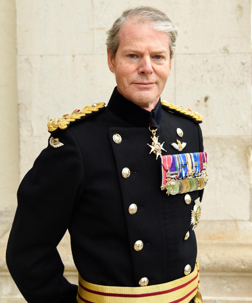 A highly decorated 4-star General of the British Army stands with hands behind his back against a white brick building