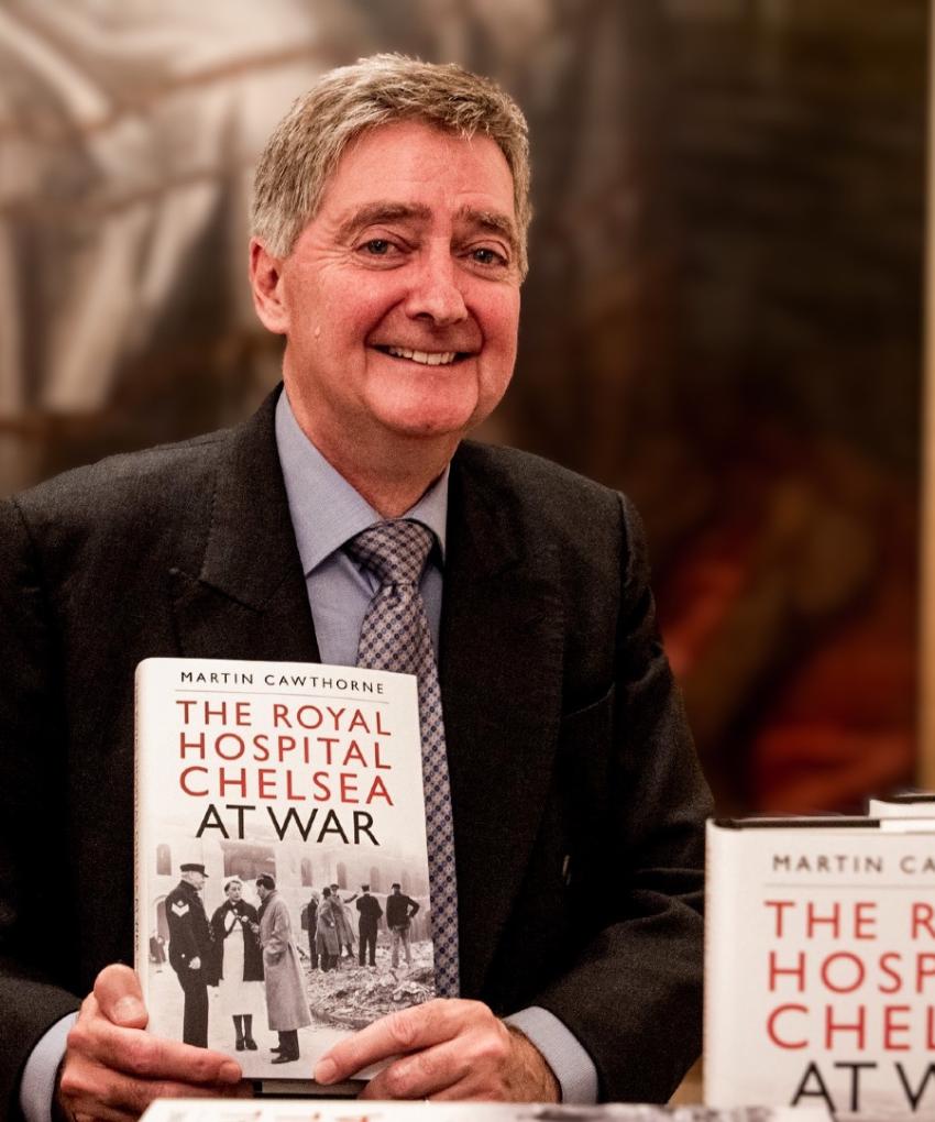 Author Martin Cawthorne is pictured wearing a suit and tie, holding a copy of his Royal Hospital Chelsea at War book, in front of a large portrait of Charles II within the State Apartments.