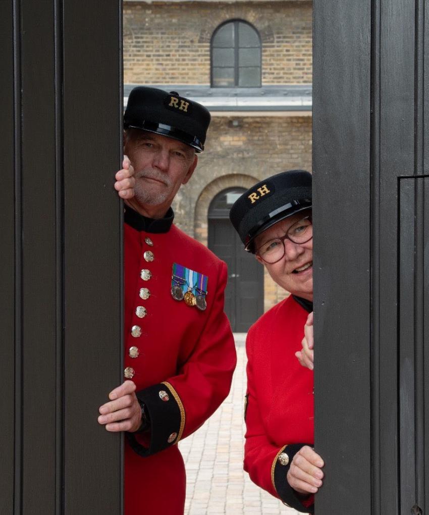Two Chelsea Pensioners, dressed in their iconic Scarlet Coats and black shako hats peek out from behind partially opened large black doors
