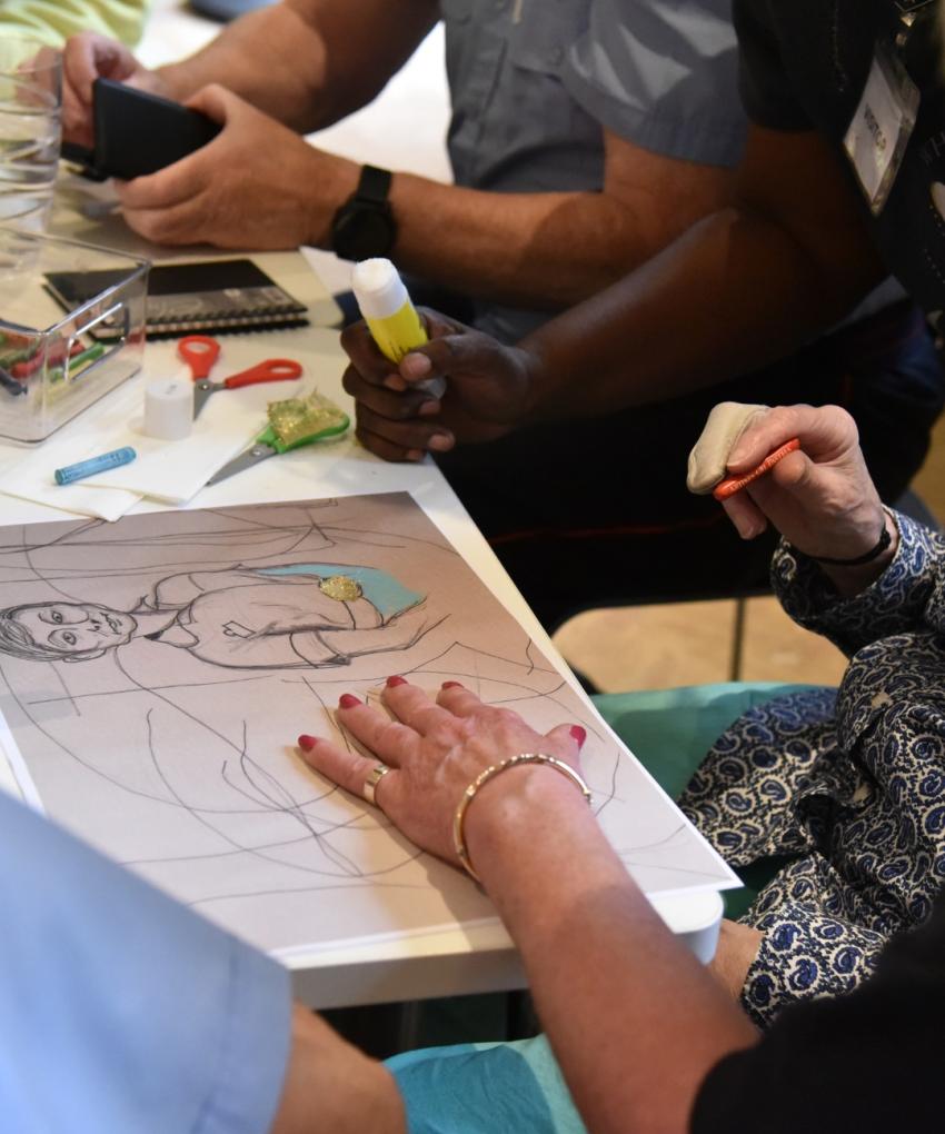 Hands of several people are shown holding various art implements, including crayons & glue. On a table are craft supplies and a sheet of paper depicting a hand-drawn sketch of a nurse.