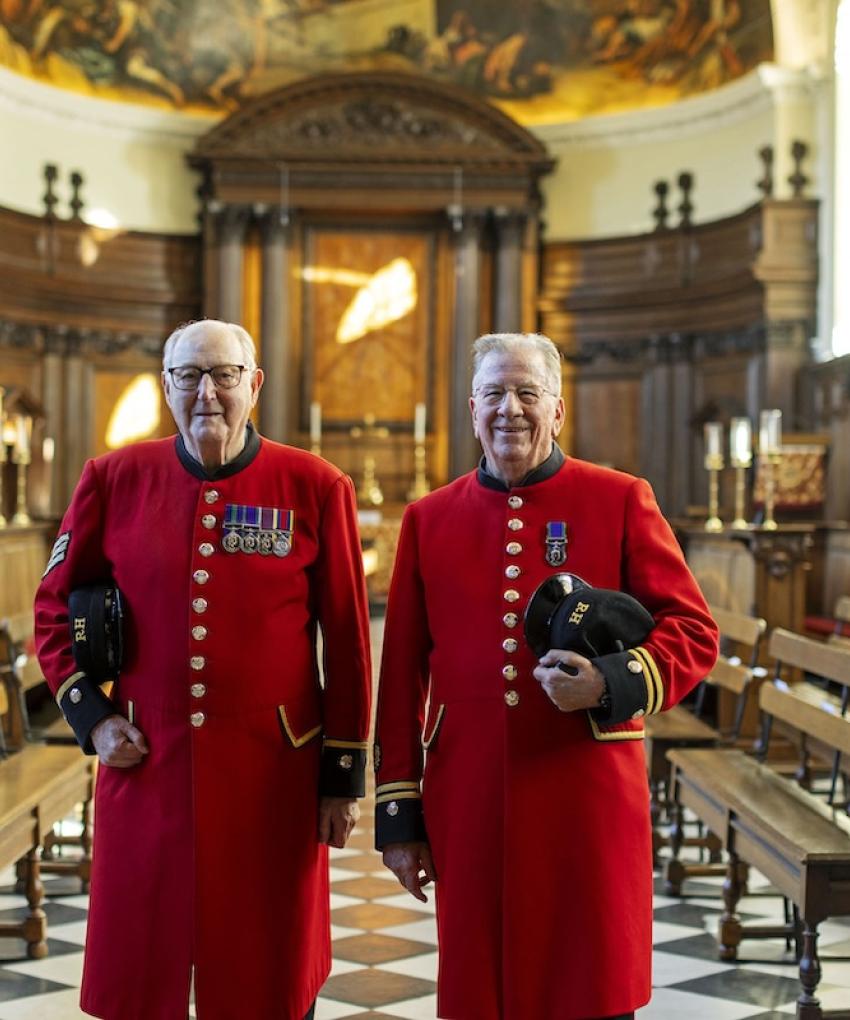 Two Chelsea Pensioners in traditional scarlet uniforms stand side by side inside the Wren Chapel at the Royal Hospital Chelsea. Wooden benches line the chapel, and the ornate altar and painted apse are visible behind them.