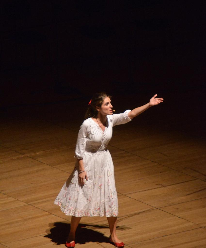 A woman in a white flowing dress stands on a wooden stage, illuminated in warm light. She raises one arm in front of her dramatically as if performing.